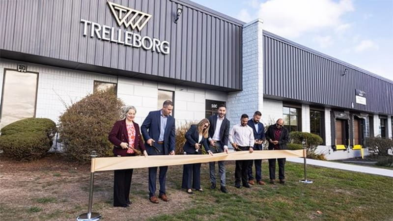 Ribbon-cutting ceremony at Trelleborg’s modern industrial facility, with seven individuals in business attire holding scissors in front of a sleek building featuring dark gray siding and large windows.