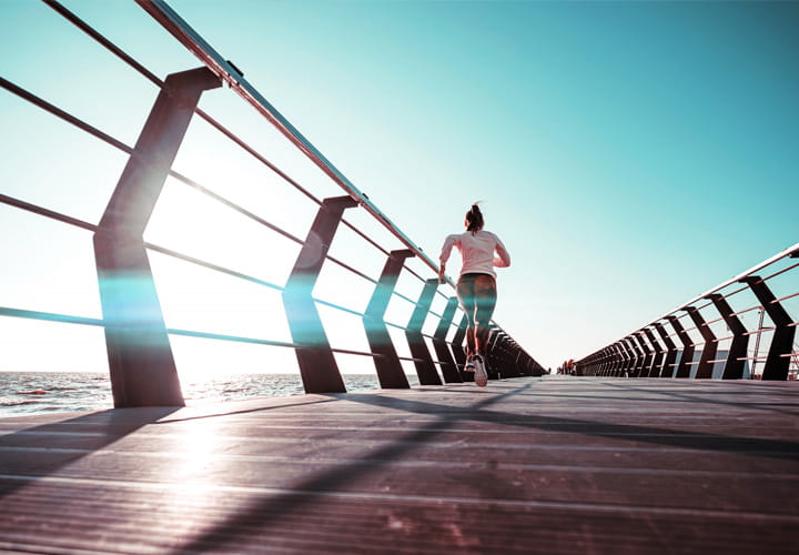 Person running along a modern boardwalk with metal railings, overlooking the ocean under a clear blue sky