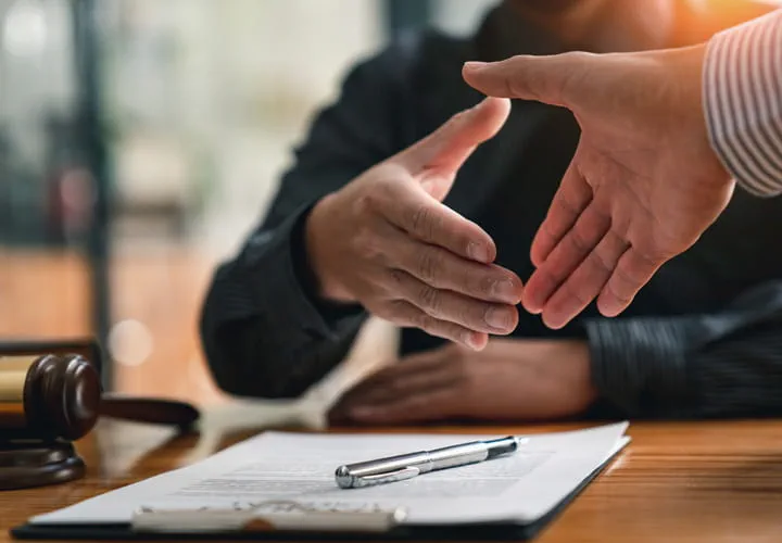 Two hands reaching for a handshake over a desk with a document and a pen