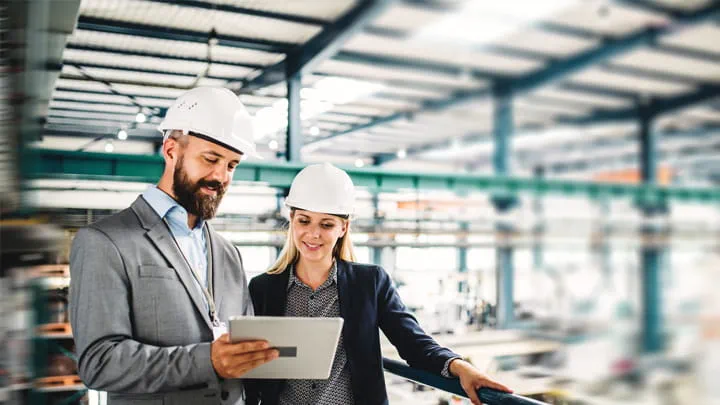 Two people wearing safety helmets reviewing information on a tablet inside an industrial facility