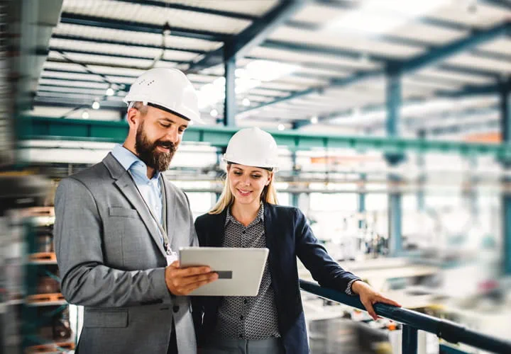 Dos personas con cascos de seguridad revisando información en una tableta dentro de una instalación industrial