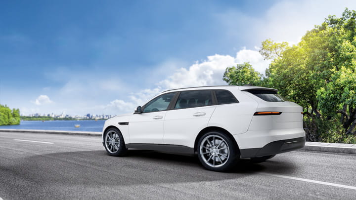 White SUV driving on a paved road with green trees and a blue sky in the background