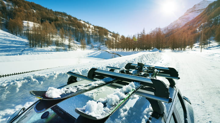 Car roof with ski equipment mounted on a roof rack in a snowy mountain landscape under bright sunlight