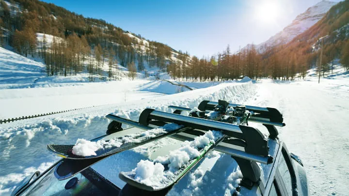 Autodach mit Skiausrüstung auf einem Dachgepäckträger in einer verschneiten Berglandschaft unter strahlendem Sonnenschein