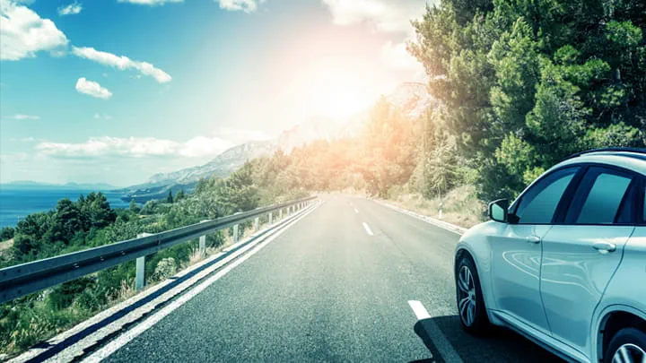 White car driving on a scenic mountain road with guardrails, surrounded by trees and hills under bright sunlight