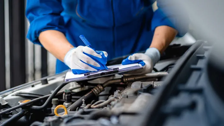 Mecánico con uniforme azul escribiendo en un portafolios mientras inspecciona el motor de un coche