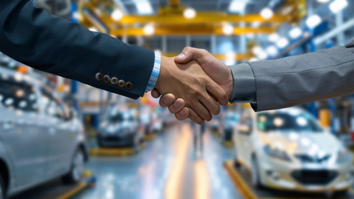 Business partners shaking hands in an automotive manufacturing facility with vehicles in the background.