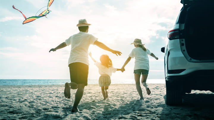 Une famille court sur une plage de sable près d'une voiture garée, avec un cerf-volant coloré qui vole dans le ciel sous un soleil radieux