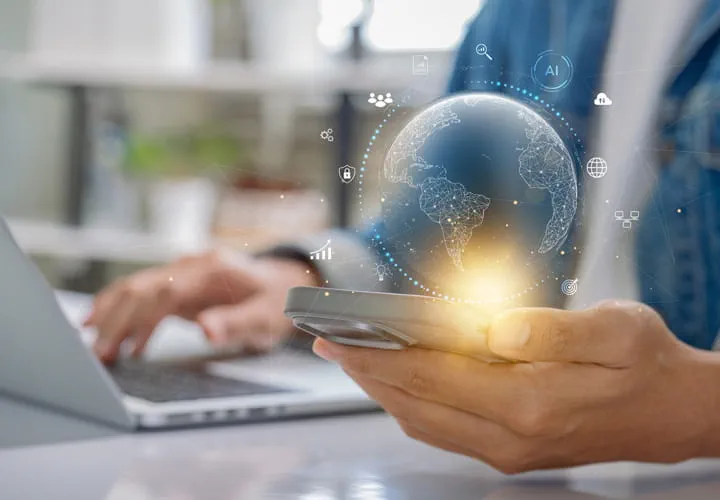 Person, die ein Smartphone mit einem leuchtenden digitalen Globus darüber in der Hand hält, sitzt an einem Schreibtisch mit einem Laptop im Hintergrund