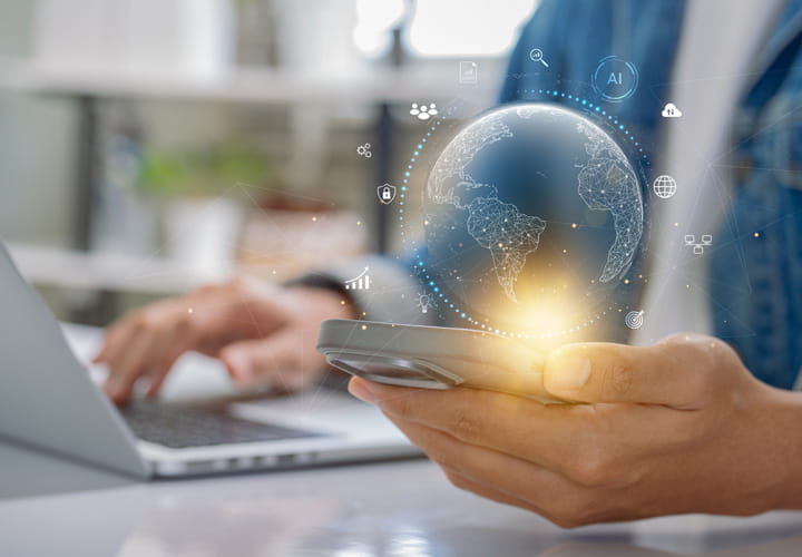 Person som håller en smartphone med en lysande digital glob ovanför, sittande vid ett skrivbord med en bärbar dator i bakgrunden