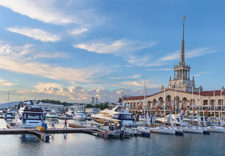 Boats and yachts docked in a marina beside a waterfront building with a tall tower under a partly cloudy sky