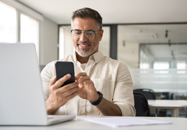 Persona sentada en un escritorio utilizando un smartphone, con un ordenador portátil abierto delante, en un entorno de oficina moderno