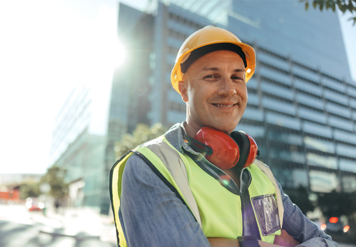 About us Happy construction worker smiling at the camera while standing with his arms crossed in the city. Mid-adult blue collar worker standing in front of high rise buildings in his workwear.